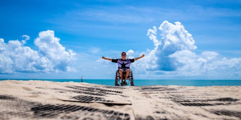 Man in wheelchair celebrating on João Pessoa beach with open arms under a clear blue sky.